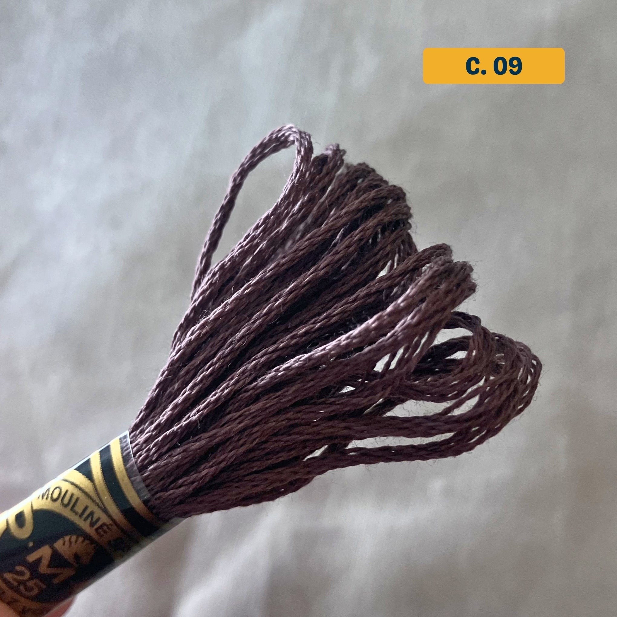 DMC 6-Strand Cotton Embroidery Floss – 09 Purple-Brown – 8m skein, 100% Egyptian cotton. Fabric background (blurred).