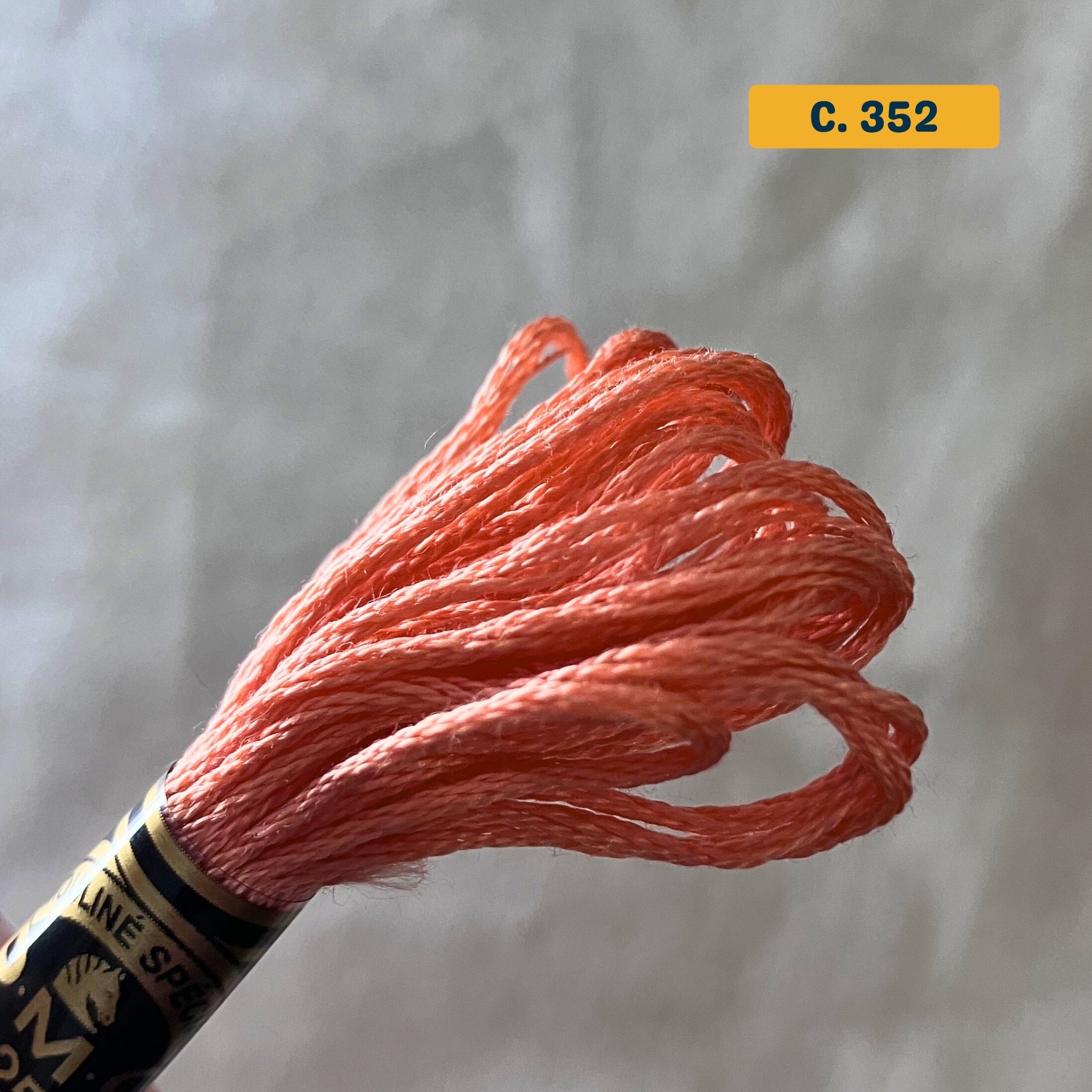 DMC 6-Strand Cotton Embroidery Floss – 352 Coral – 8m skein, 100% Egyptian cotton. Fabric background (blurred).