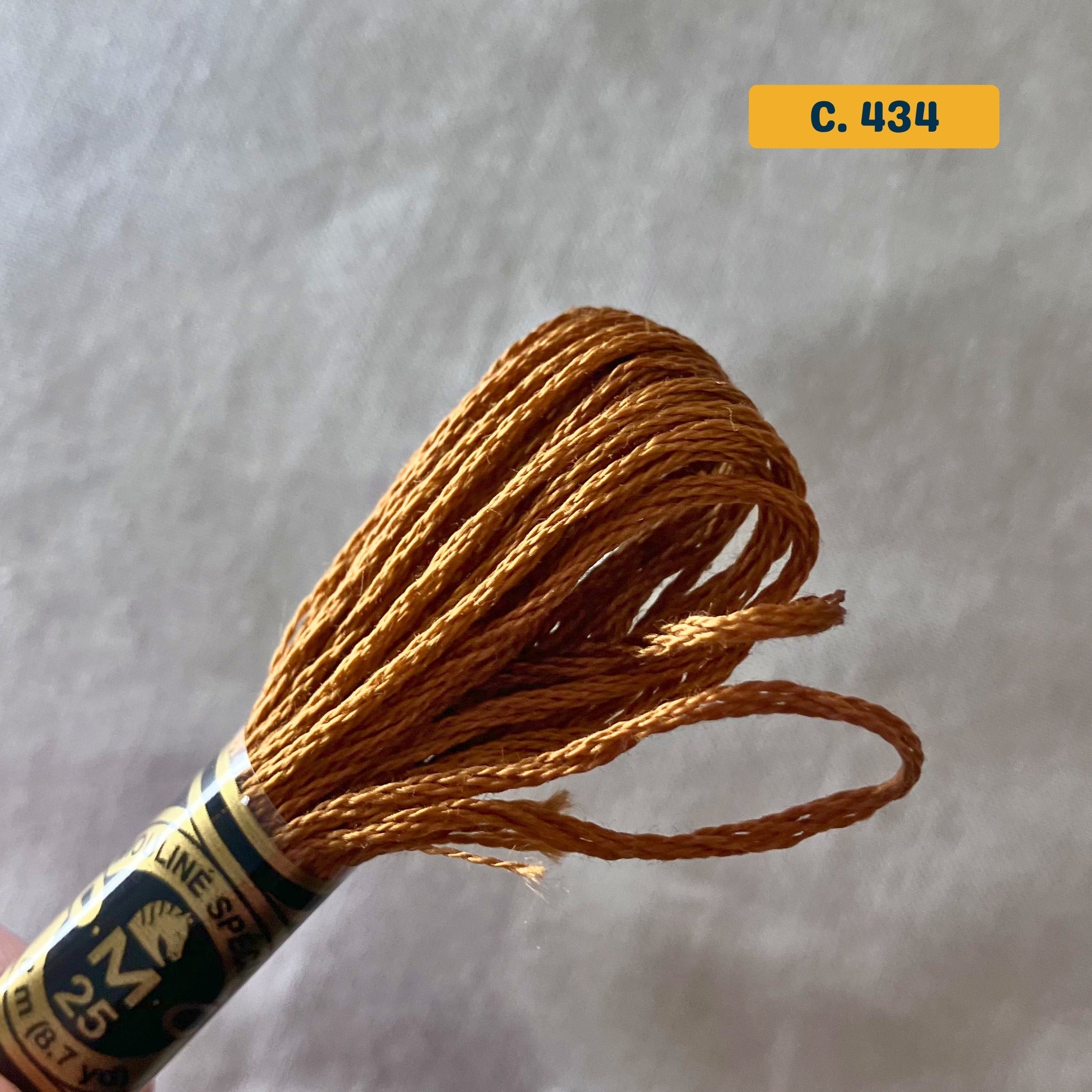 DMC 6-Strand Cotton Embroidery Floss – 434 Bronze – 8m skein, 100% Egyptian cotton. Fabric background (blurred).