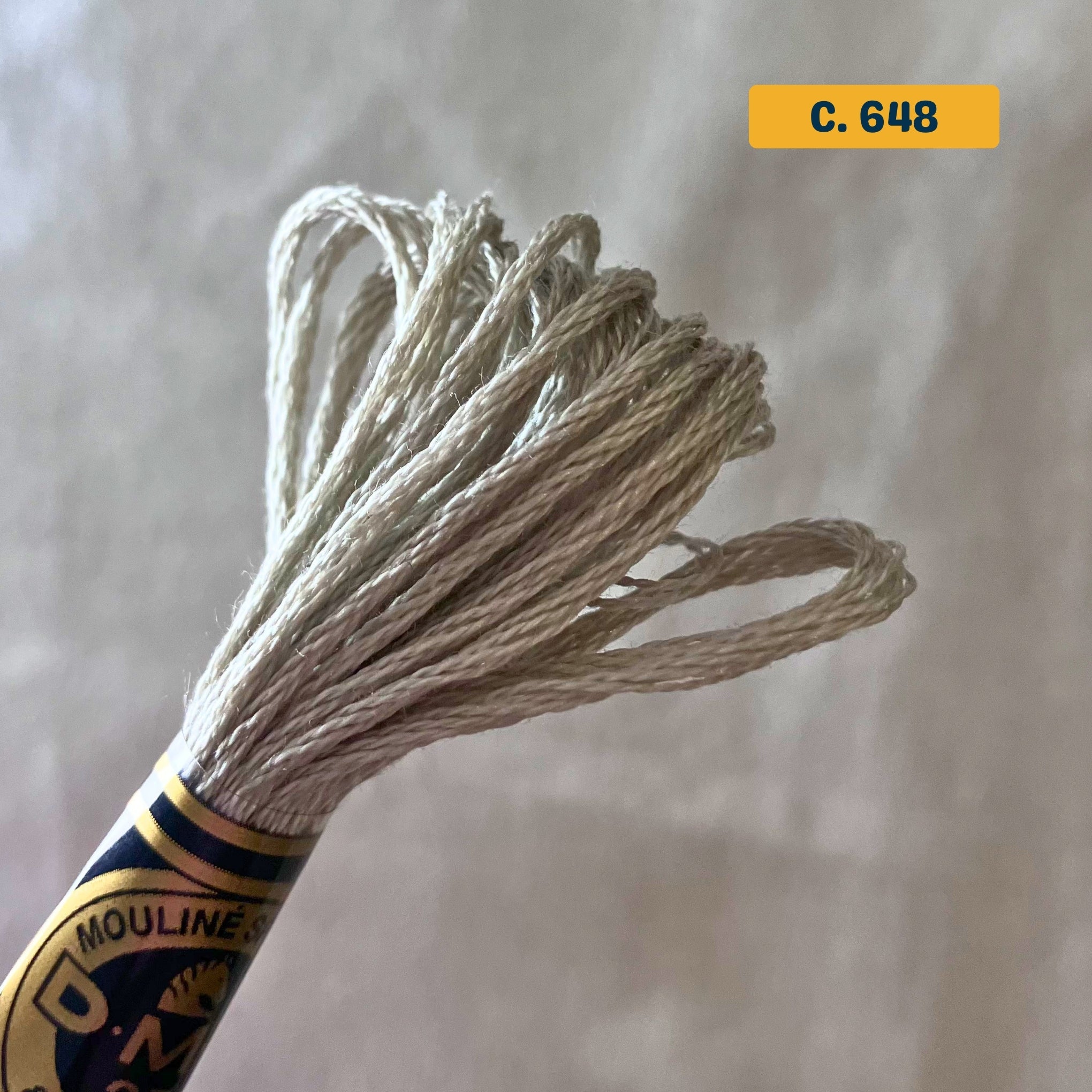 DMC 6-Strand Cotton Embroidery Floss – 648 Light Grey – 8m skein, 100% Egyptian cotton. Fabric background (blurred).