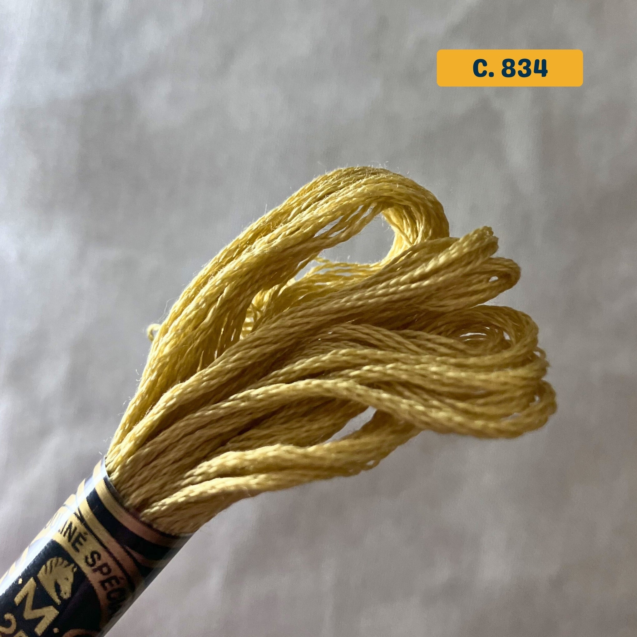 DMC 6-Strand Cotton Embroidery Floss – 834 Mustard – 8m skein, 100% Egyptian cotton. Fabric background (blurred).