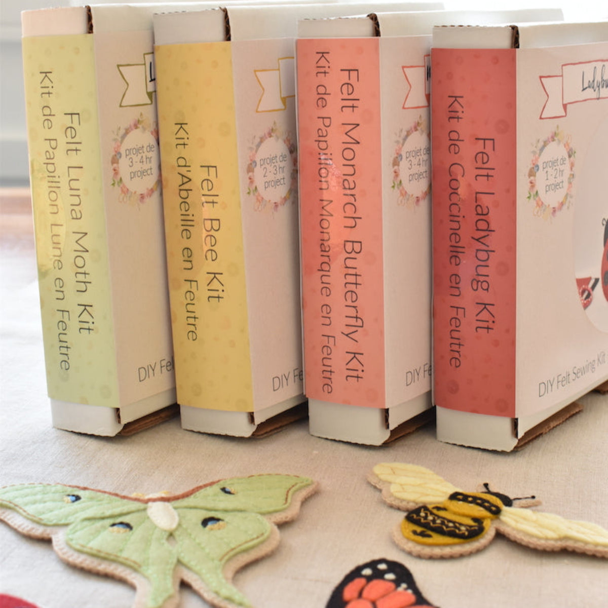 Four DIY craft kits in pastel-coloured cardboard boxes labeled for Felt Insect Craft Kits, arranged on a light wooden surface with insect-shaped felt in the foreground.