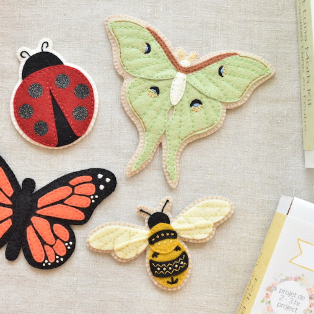 Four embroidered fabric butterfly and insect patches arranged on a light beige textured surface, with partial books visible around the edges. The patches include a red ladybug, a pale green moth, an orange-and-black butterfly, and a yellow-and-black bee.