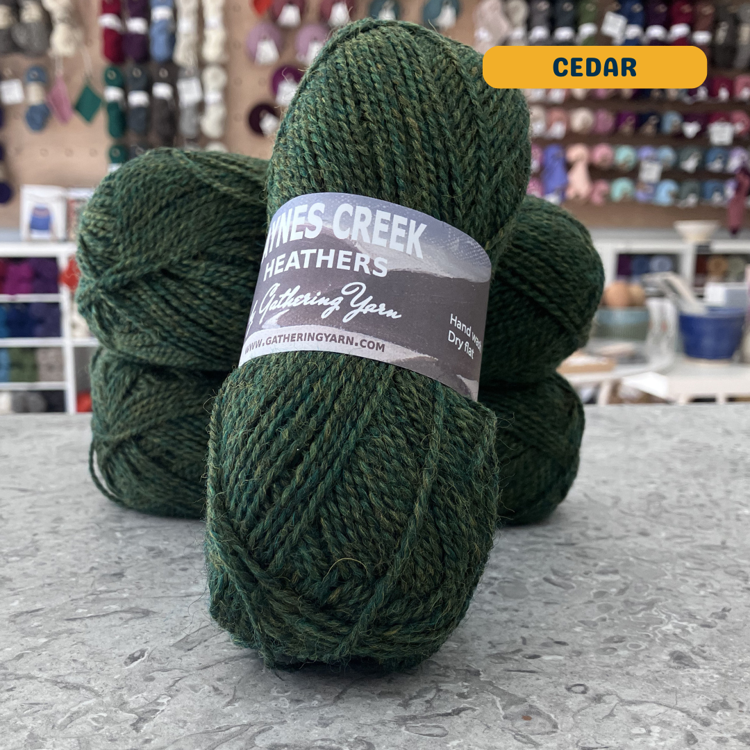 50g dark green (cedar) Peruvian highland wool DK yarn, 'Haynes Creek Heathers by Gathering Yarn' label visible, craft shop background (blurred)

