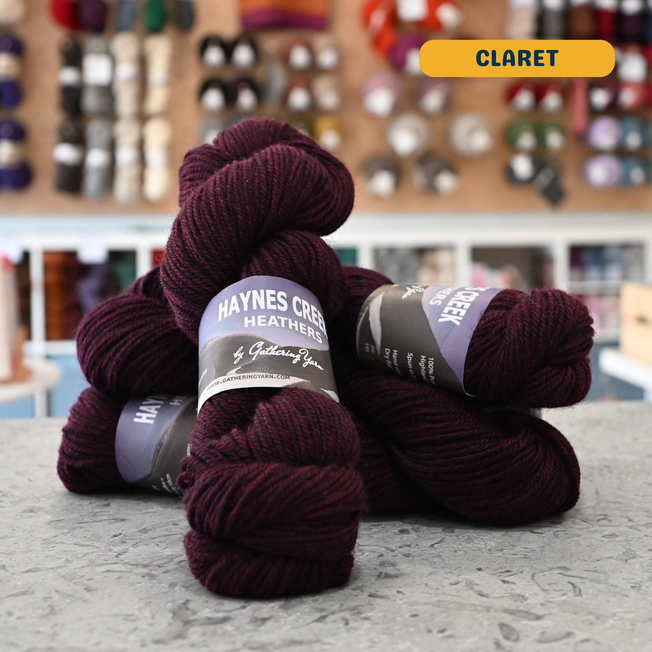 100g dark purple (claret) Peruvian highland wool worsted skeins, 'Haynes Creek Heathers by Gathering Yarn' label visible, craft shop background (blurred)
