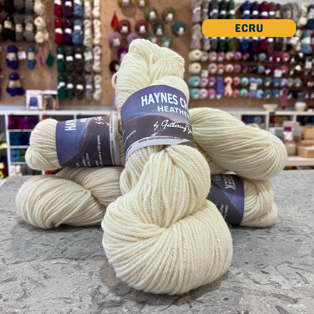 100g natural white (ecru) Peruvian highland wool worsted skeins, 'Haynes Creek Heathers by Gathering Yarn' label visible, craft shop background (blurred)