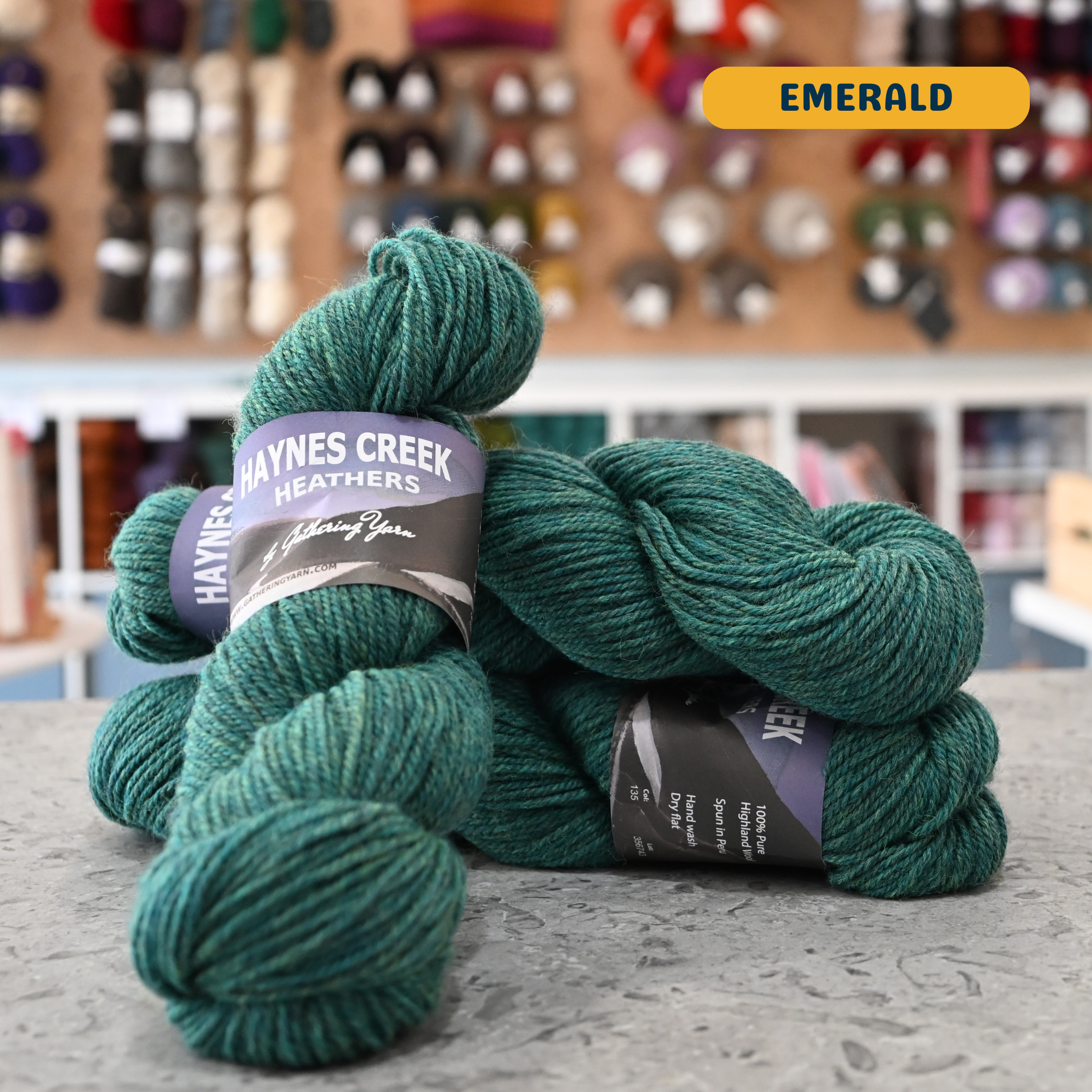 100g green (emerald) Peruvian highland wool worsted skeins, 'Haynes Creek Heathers by Gathering Yarn' label visible, craft shop background (blurred)