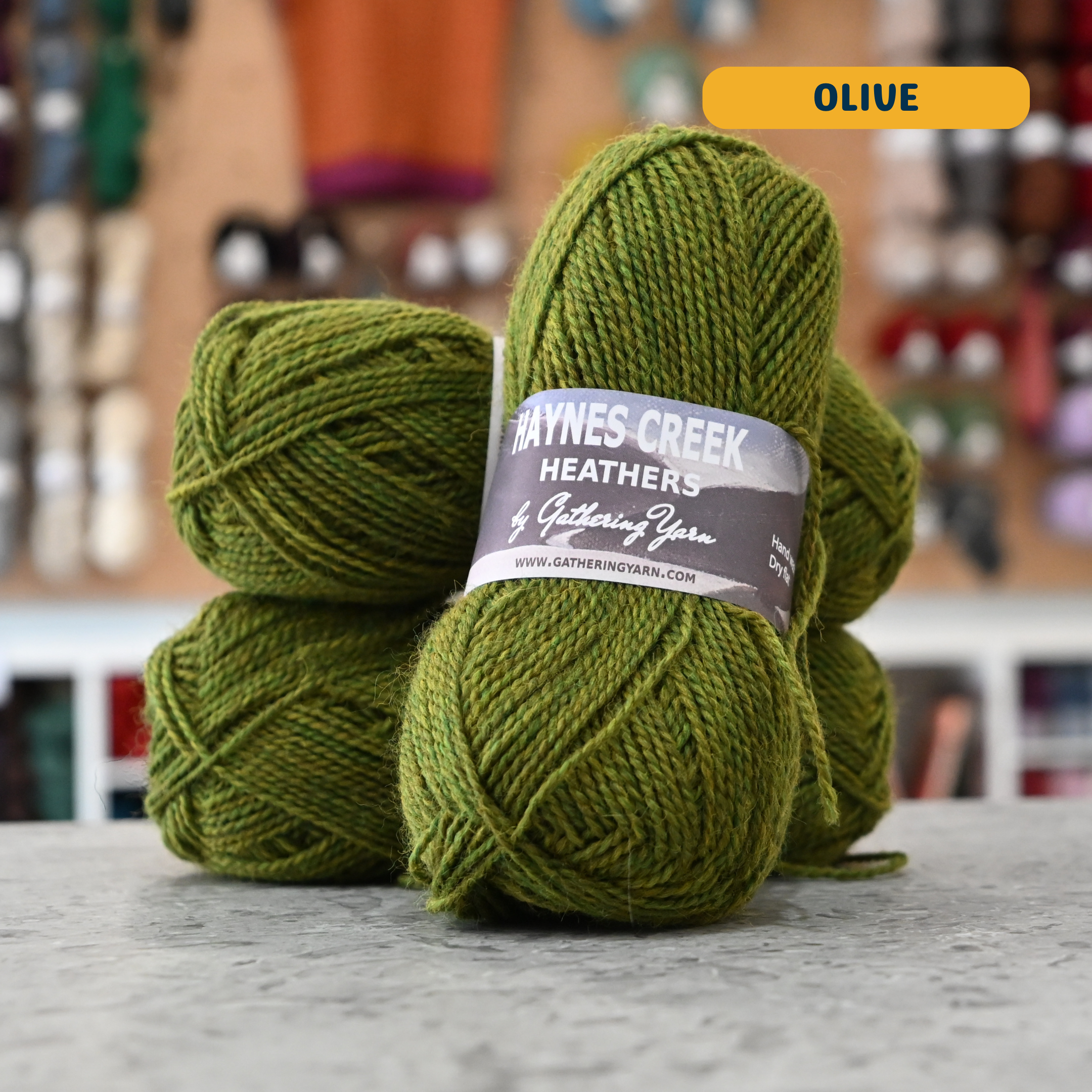 50g green (olive) Peruvian highland wool DK yarn, 'Haynes Creek Heathers by Gathering Yarn' label visible, craft shop background (blurred)
