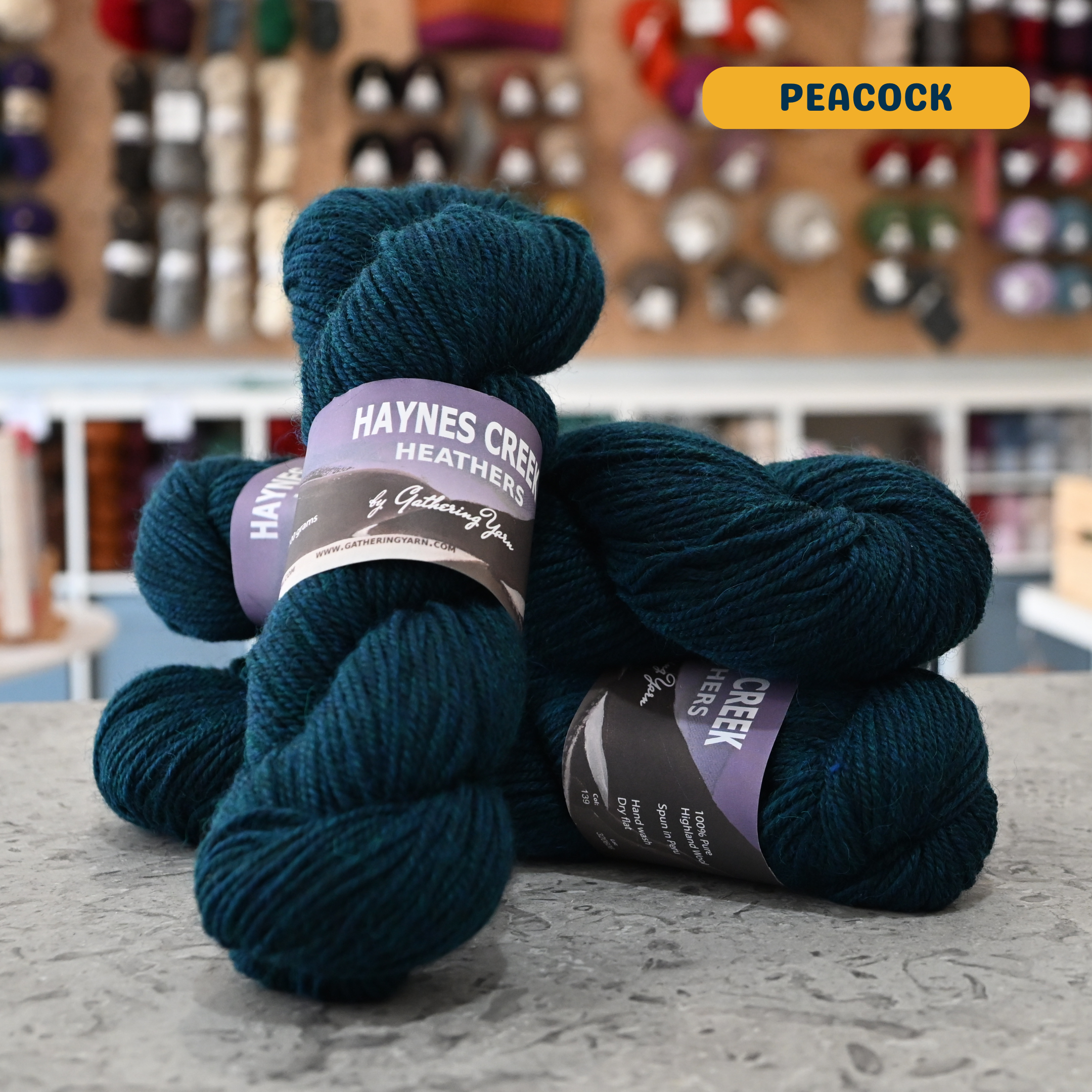 100g dark blue (peacock) Peruvian highland wool worsted skeins, 'Haynes Creek Heathers by Gathering Yarn' label visible, craft shop background (blurred)