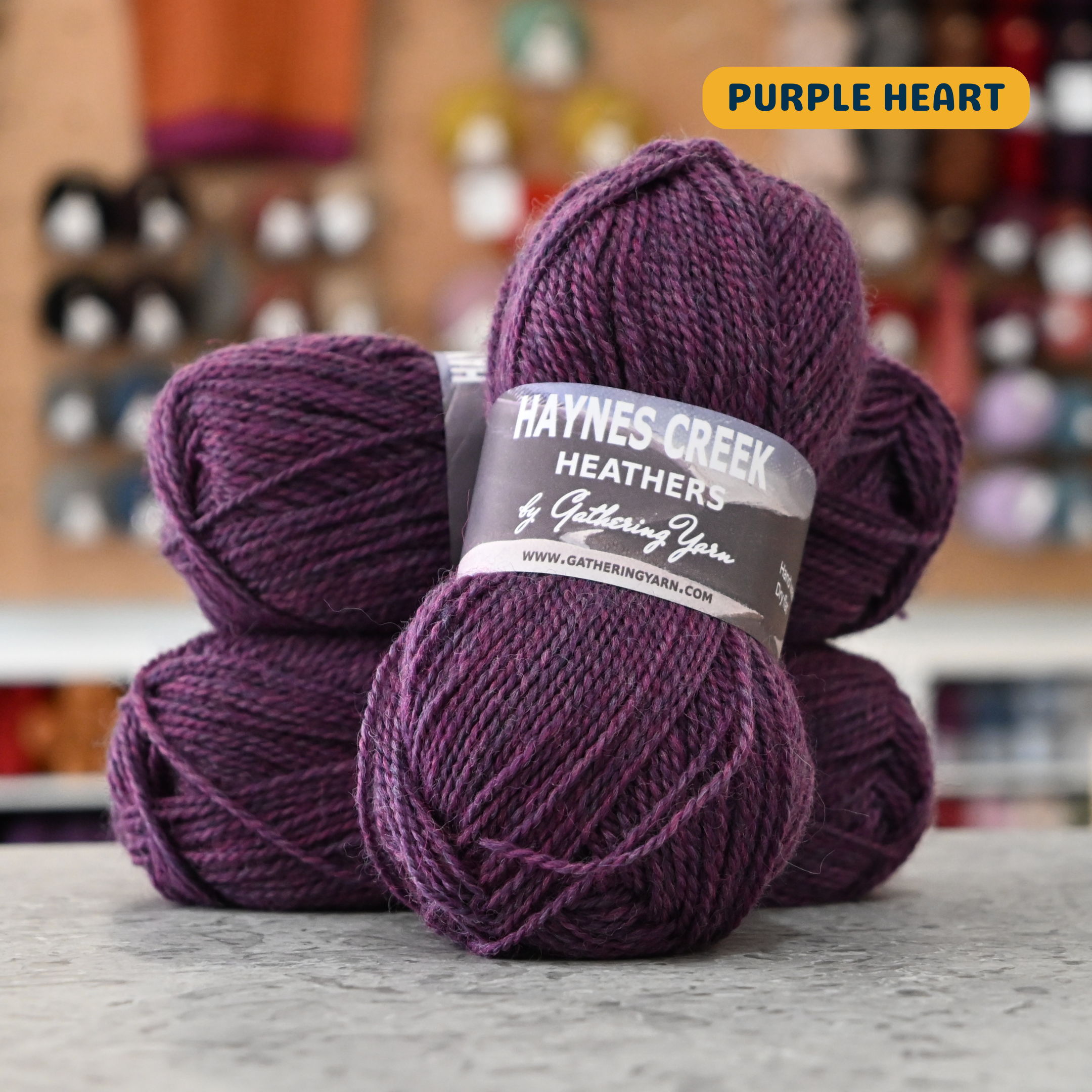 50g dark purple (purple heart) Peruvian highland wool DK yarn, 'Haynes Creek Heathers by Gathering Yarn' label visible, craft shop background (blurred)