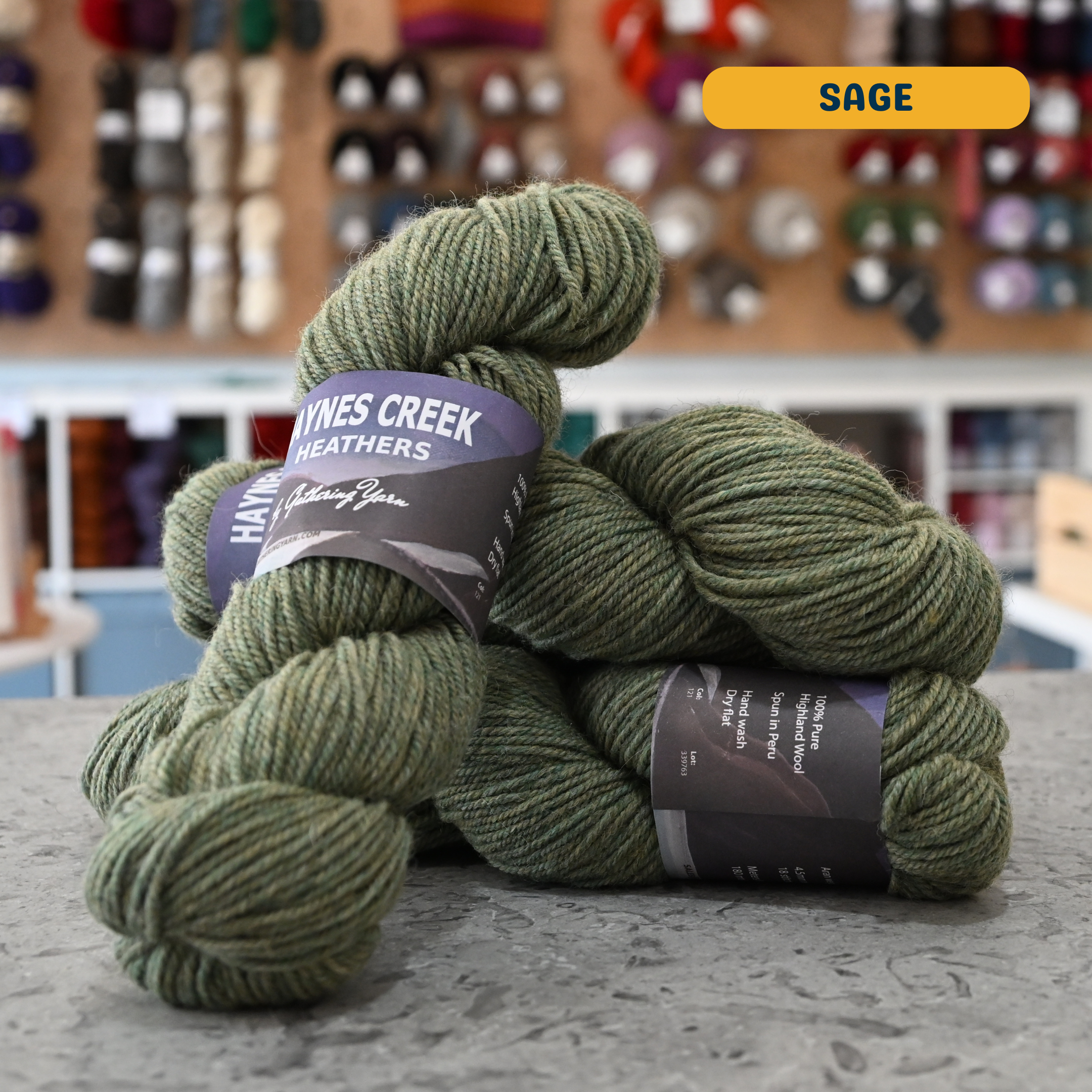 100g sage green Peruvian highland wool worsted skeins, 'Haynes Creek Heathers by Gathering Yarn' label visible, craft shop background (blurred)