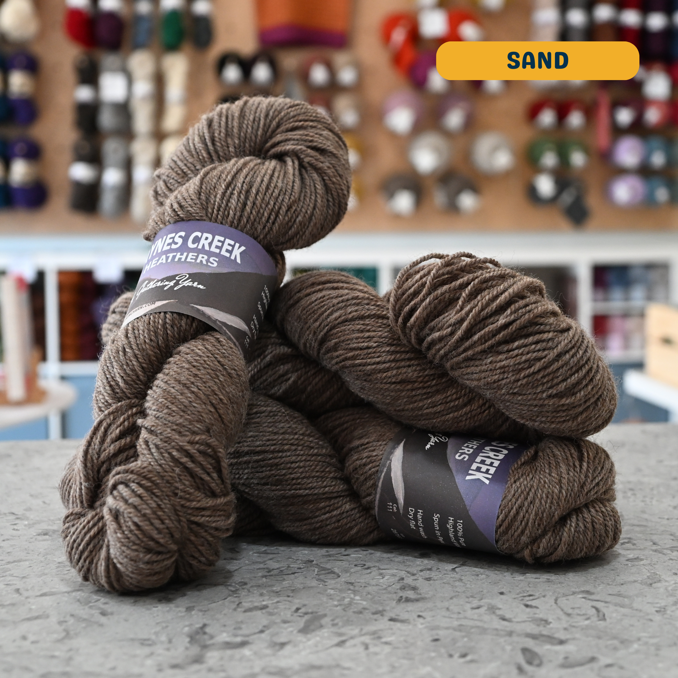 100g beige-brown (sand) Peruvian highland wool worsted skeins, 'Haynes Creek Heathers by Gathering Yarn' label visible, craft shop background (blurred)