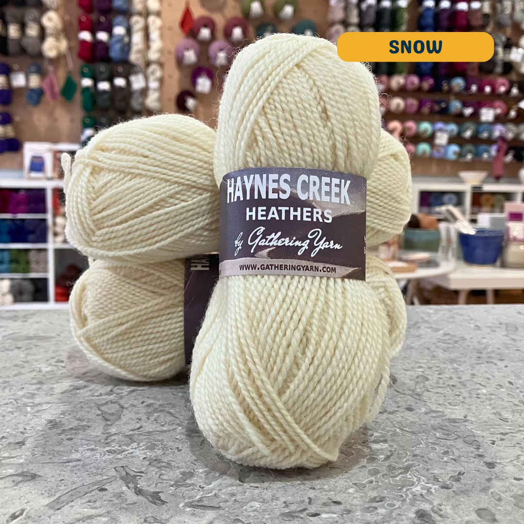 50g natural white (snow) Peruvian highland wool DK yarn, 'Haynes Creek Heathers by Gathering Yarn' label visible, craft shop background (blurred)