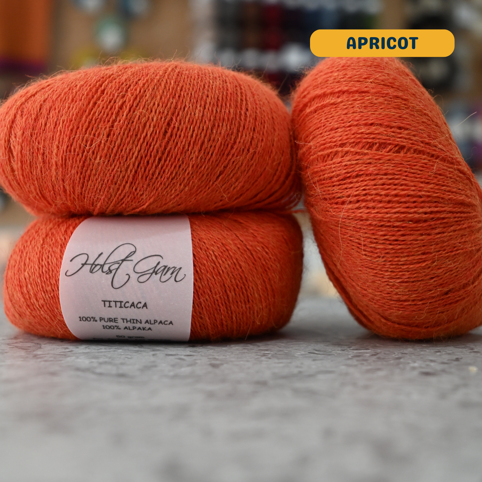 50g bright orange (apricot) alpaca lace yarn, 'Holst Garn Titicaca 100% Pure Thin Alpaca' label visible, craft shop background (blurred)