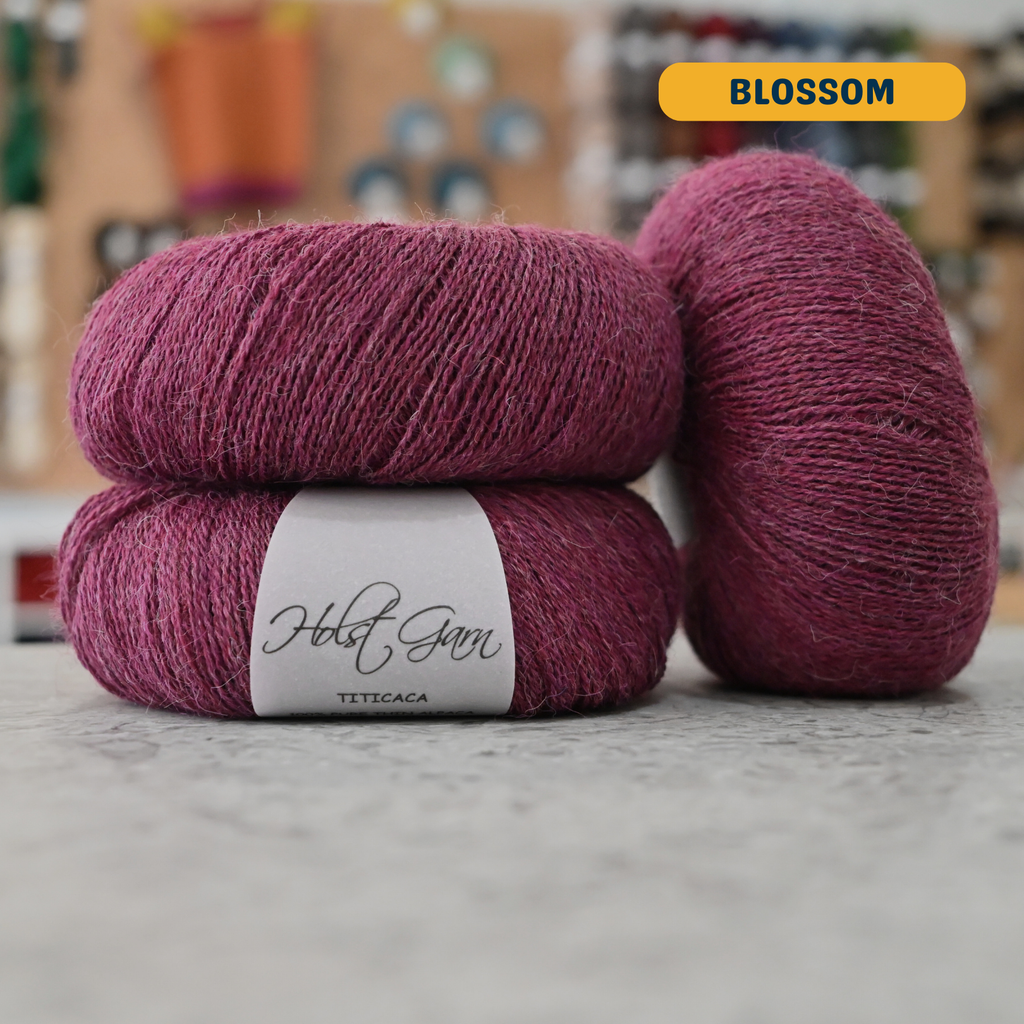 50g red-purple (blossom) alpaca lace yarn, 'Holst Garn Titicaca 100% Pure Thin Alpaca' label visible, craft shop background (blurred)