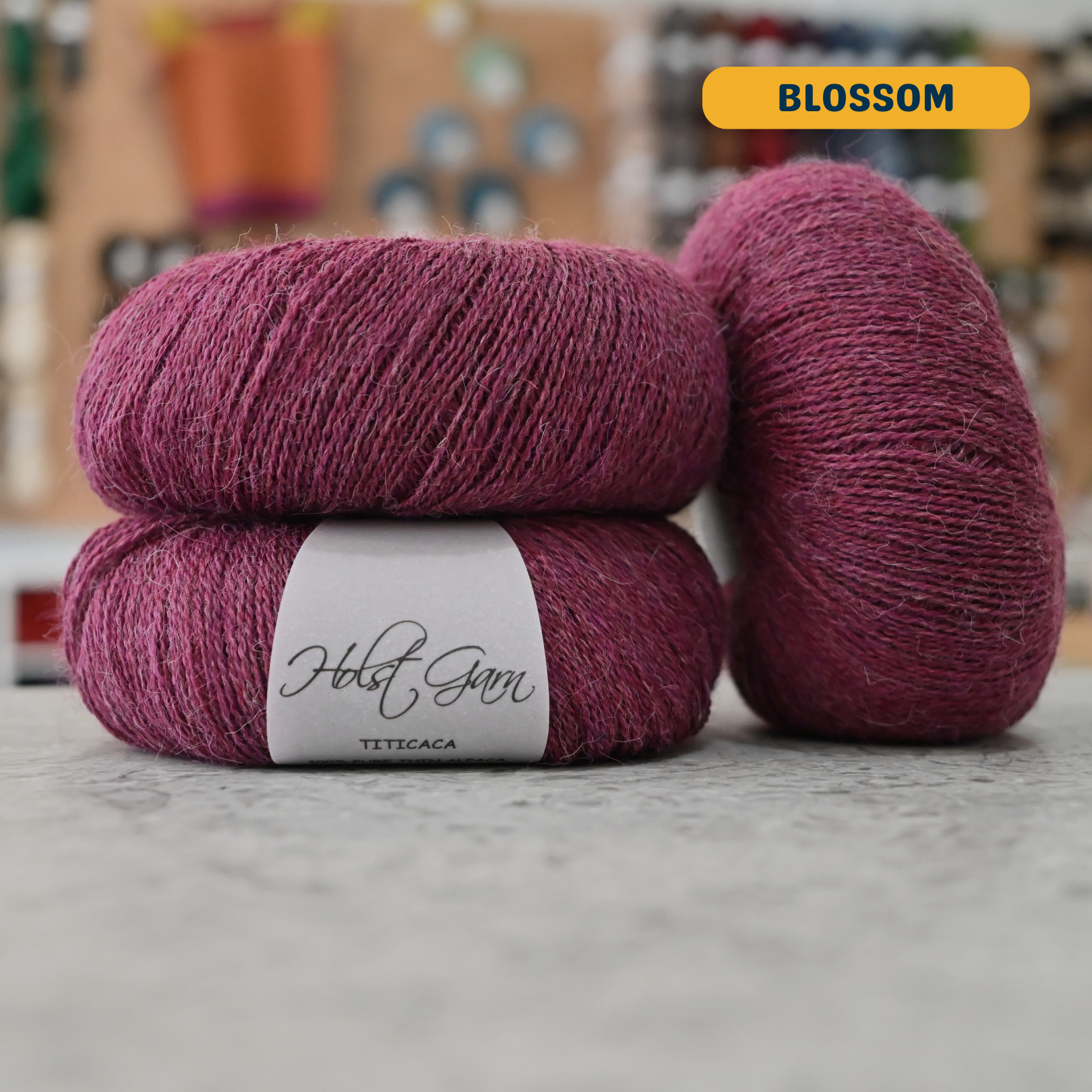 50g red-purple (blossom) alpaca lace yarn, 'Holst Garn Titicaca 100% Pure Thin Alpaca' label visible, craft shop background (blurred)