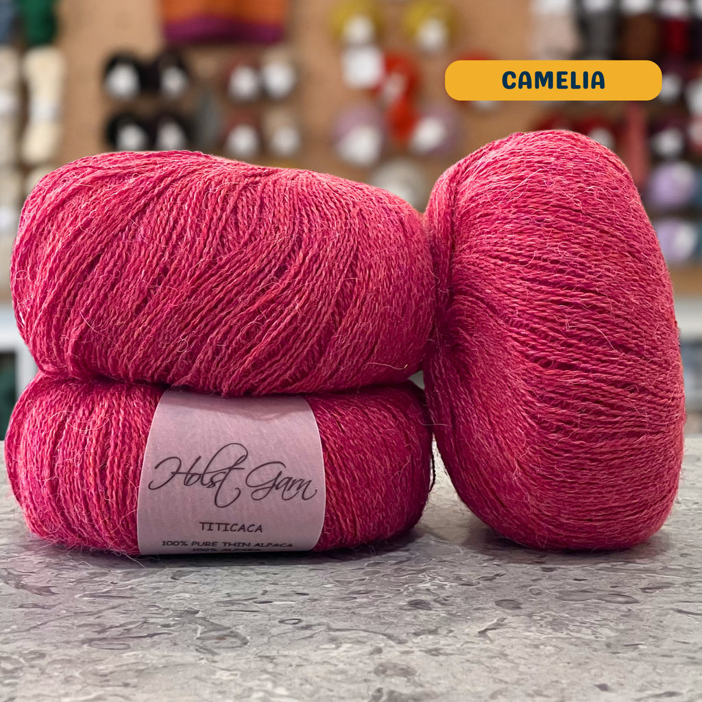 50g pink-coral (camelia) alpaca lace yarn, 'Holst Garn Titicaca 100% Pure Thin Alpaca' label visible, craft shop background (blurred)