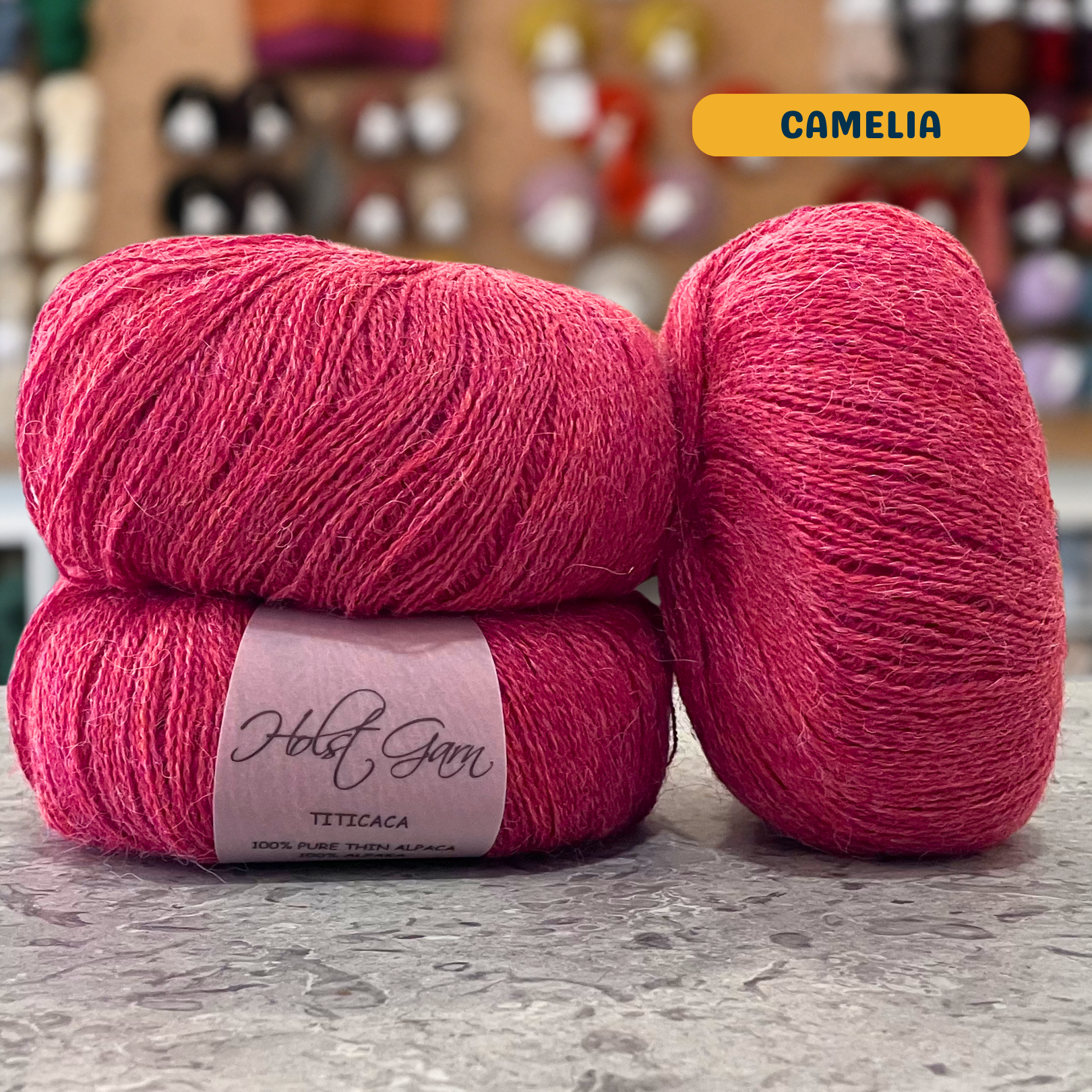 50g pink-coral (camelia) alpaca lace yarn, 'Holst Garn Titicaca 100% Pure Thin Alpaca' label visible, craft shop background (blurred)