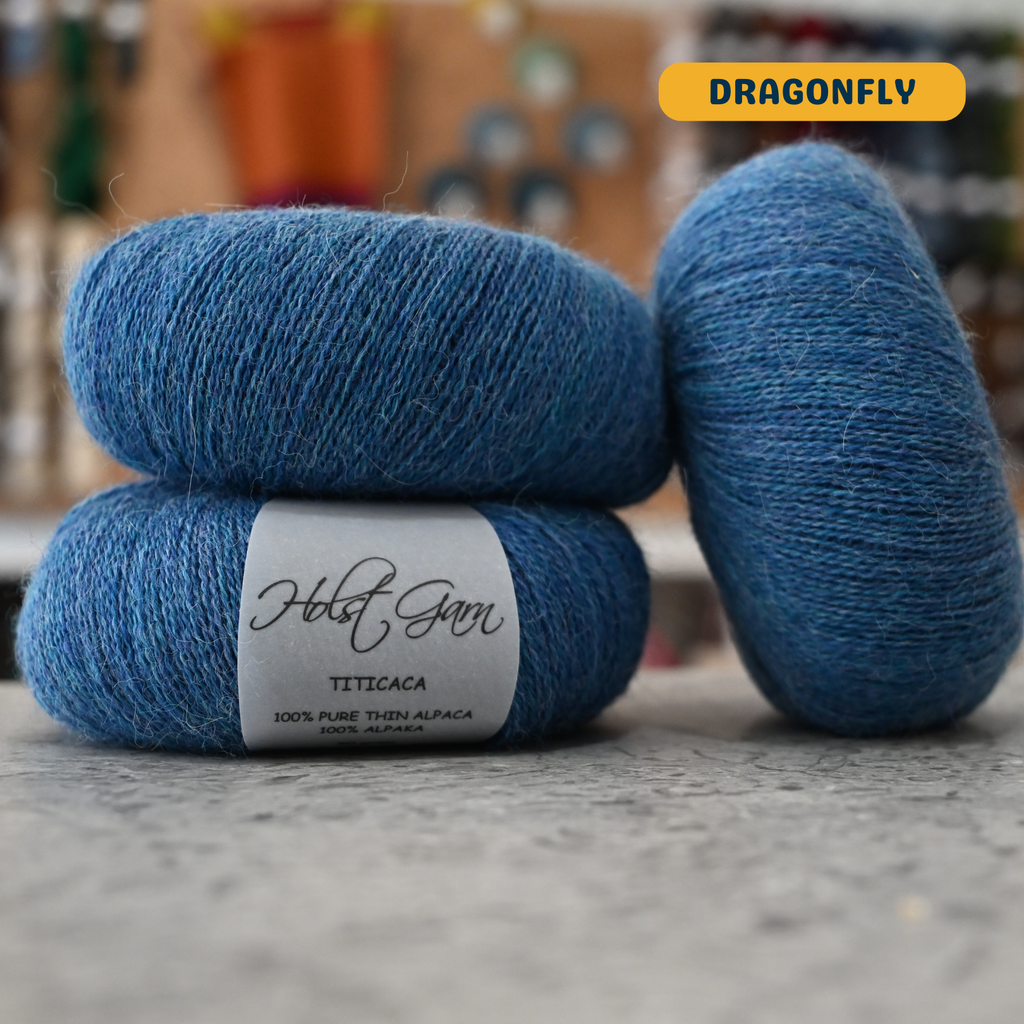 50g blue (dragonfly) alpaca lace yarn, 'Holst Garn Titicaca 100% Pure Thin Alpaca' label visible, craft shop background (blurred)