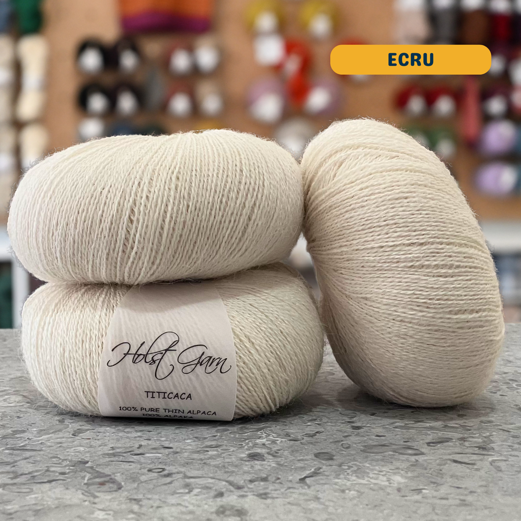 50g natural white (ecru) alpaca lace yarn, 'Holst Garn Titicaca 100% Pure Thin Alpaca' label visible, craft shop background (blurred)