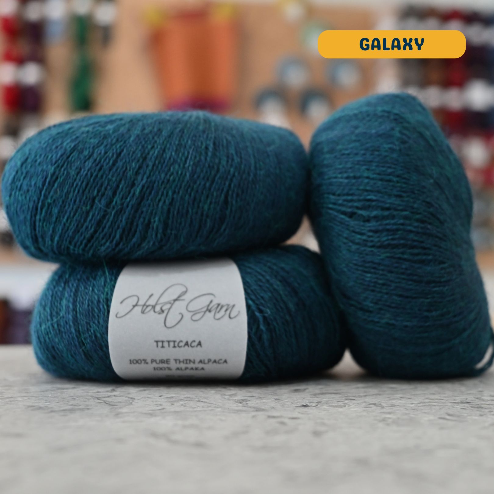 50g dark teal (galaxy) alpaca lace yarn, 'Holst Garn Titicaca 100% Pure Thin Alpaca' label visible, craft shop background (blurred)