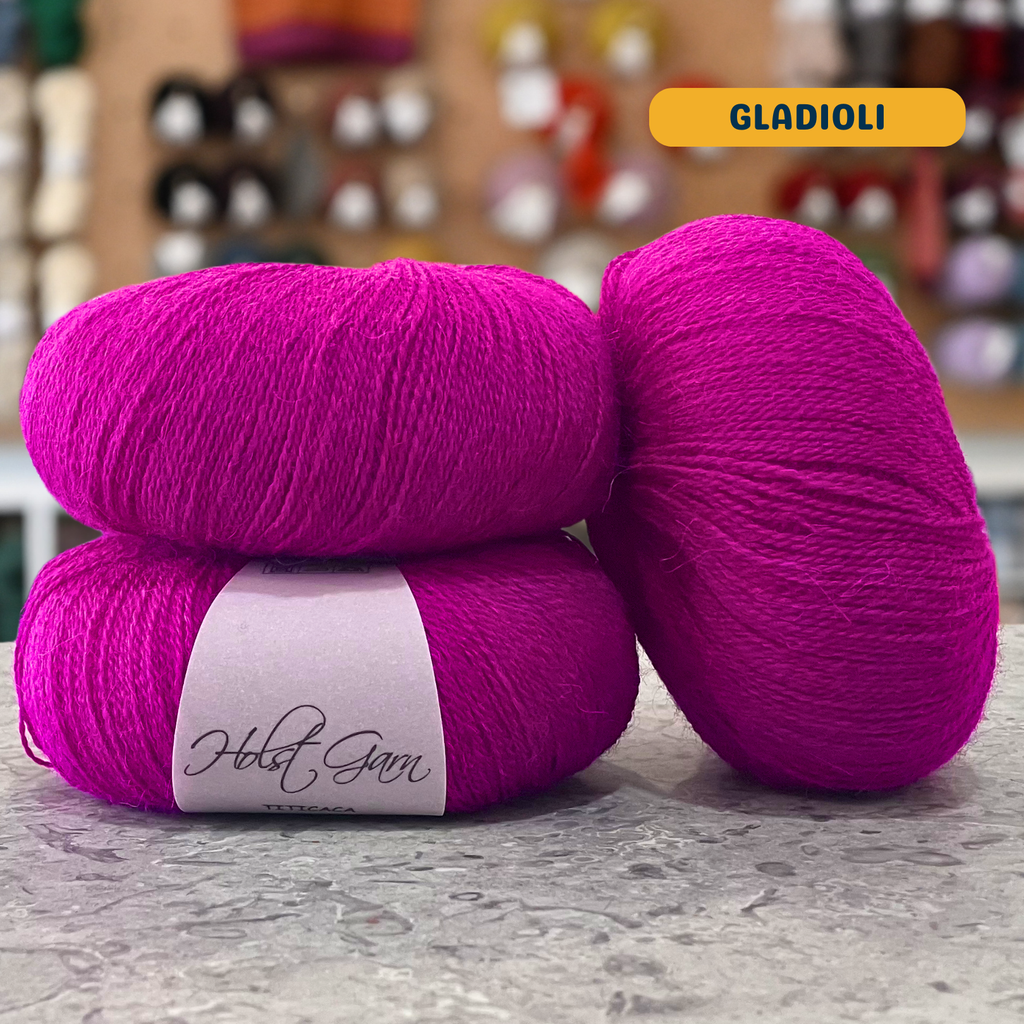 50g vibrant pink (gladioli) alpaca lace yarn, 'Holst Garn Titicaca 100% Pure Thin Alpaca' label visible, craft shop background (blurred)