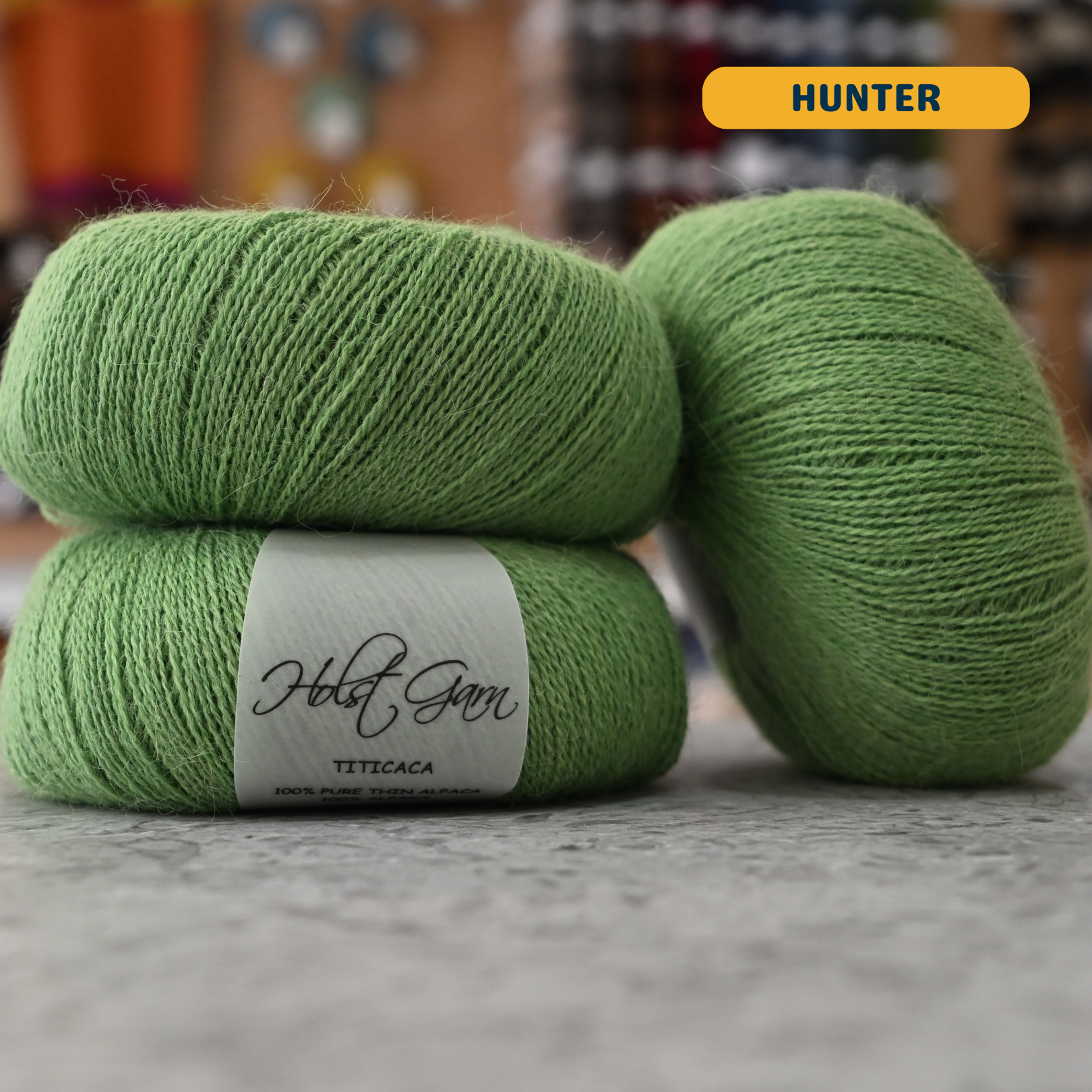 50g green (hunter) alpaca lace yarn, 'Holst Garn Titicaca 100% Pure Thin Alpaca' label visible, craft shop background (blurred)