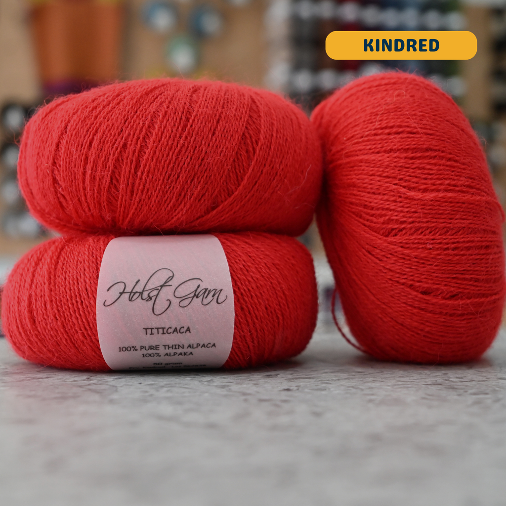 50g bright red (kindred) alpaca lace yarn, 'Holst Garn Titicaca 100% Pure Thin Alpaca' label visible, craft shop background (blurred)