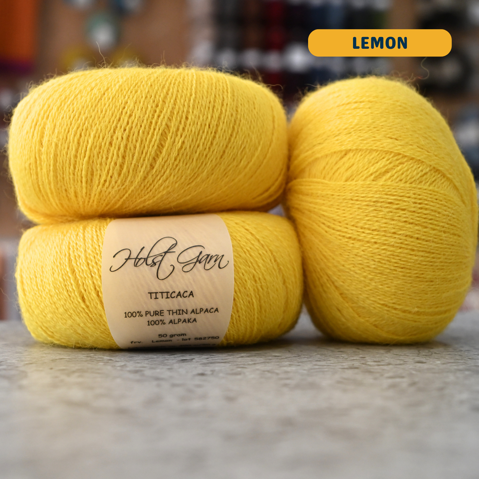 50g bright yellow (lemon) alpaca lace yarn, 'Holst Garn Titicaca 100% Pure Thin Alpaca' label visible, craft shop background (blurred)
