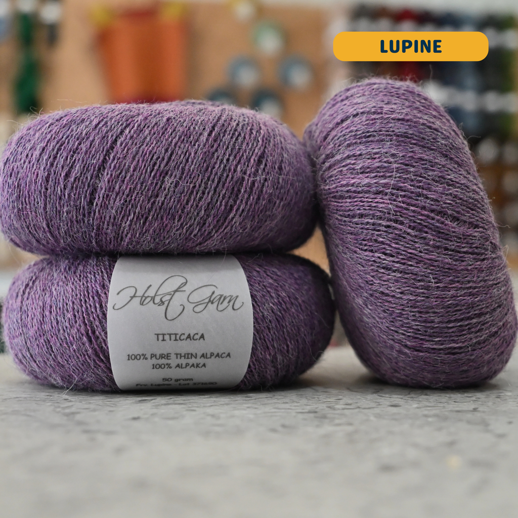 50g purple (lupine) alpaca lace yarn, 'Holst Garn Titicaca 100% Pure Thin Alpaca' label visible, craft shop background (blurred)