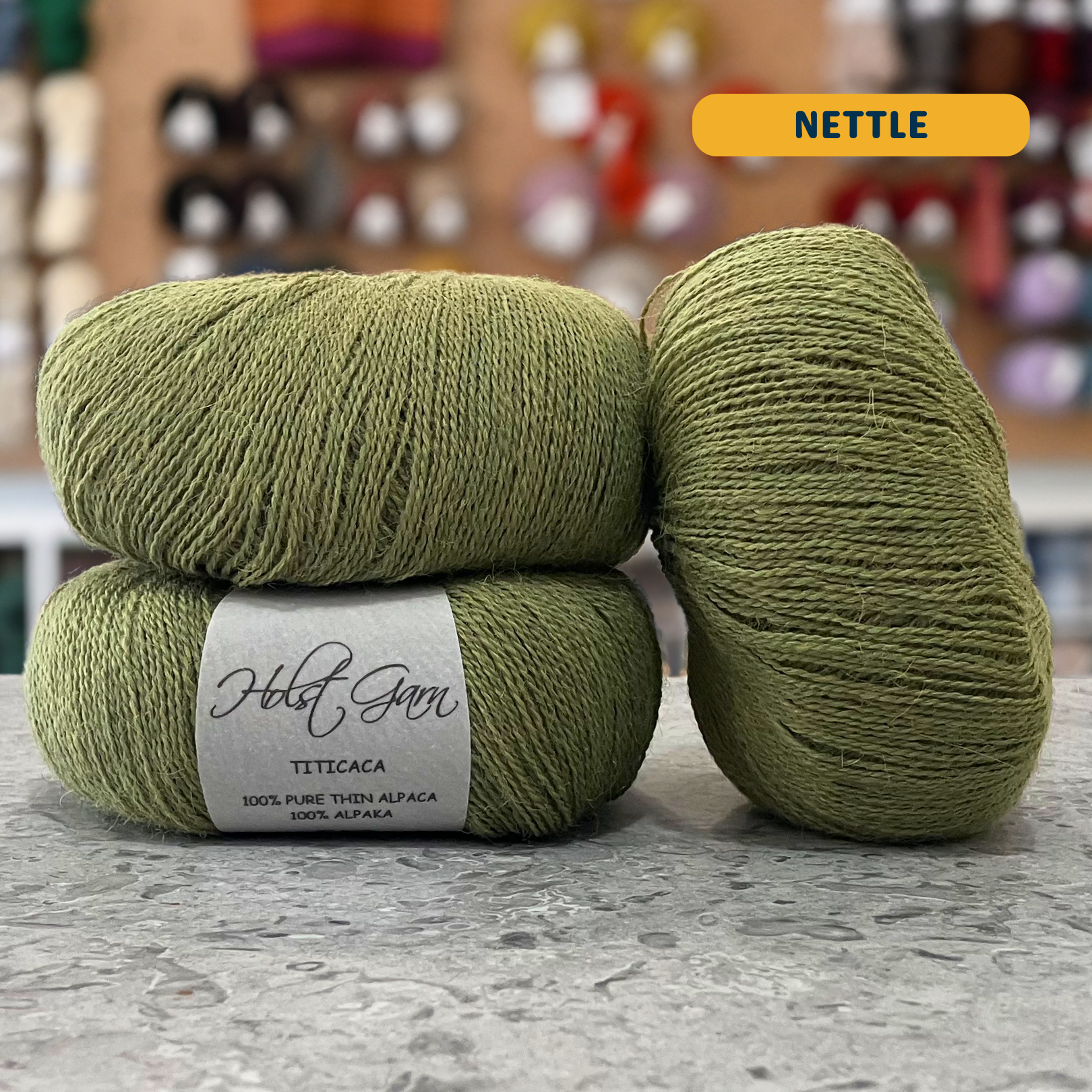 50g green (nettle) alpaca lace yarn, 'Holst Garn Titicaca 100% Pure Thin Alpaca' label visible, craft shop background (blurred)