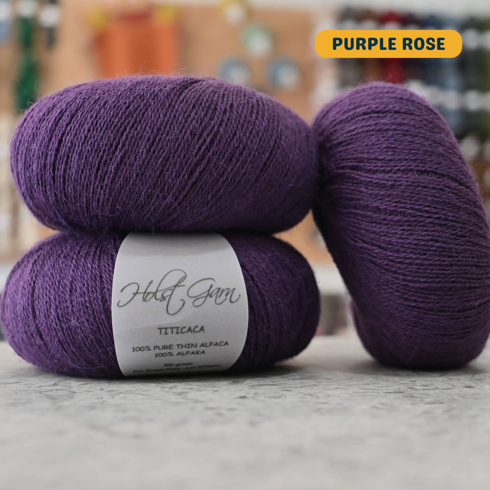 50g dark purple (purple rose) alpaca lace yarn, 'Holst Garn Titicaca 100% Pure Thin Alpaca' label visible, craft shop background (blurred)