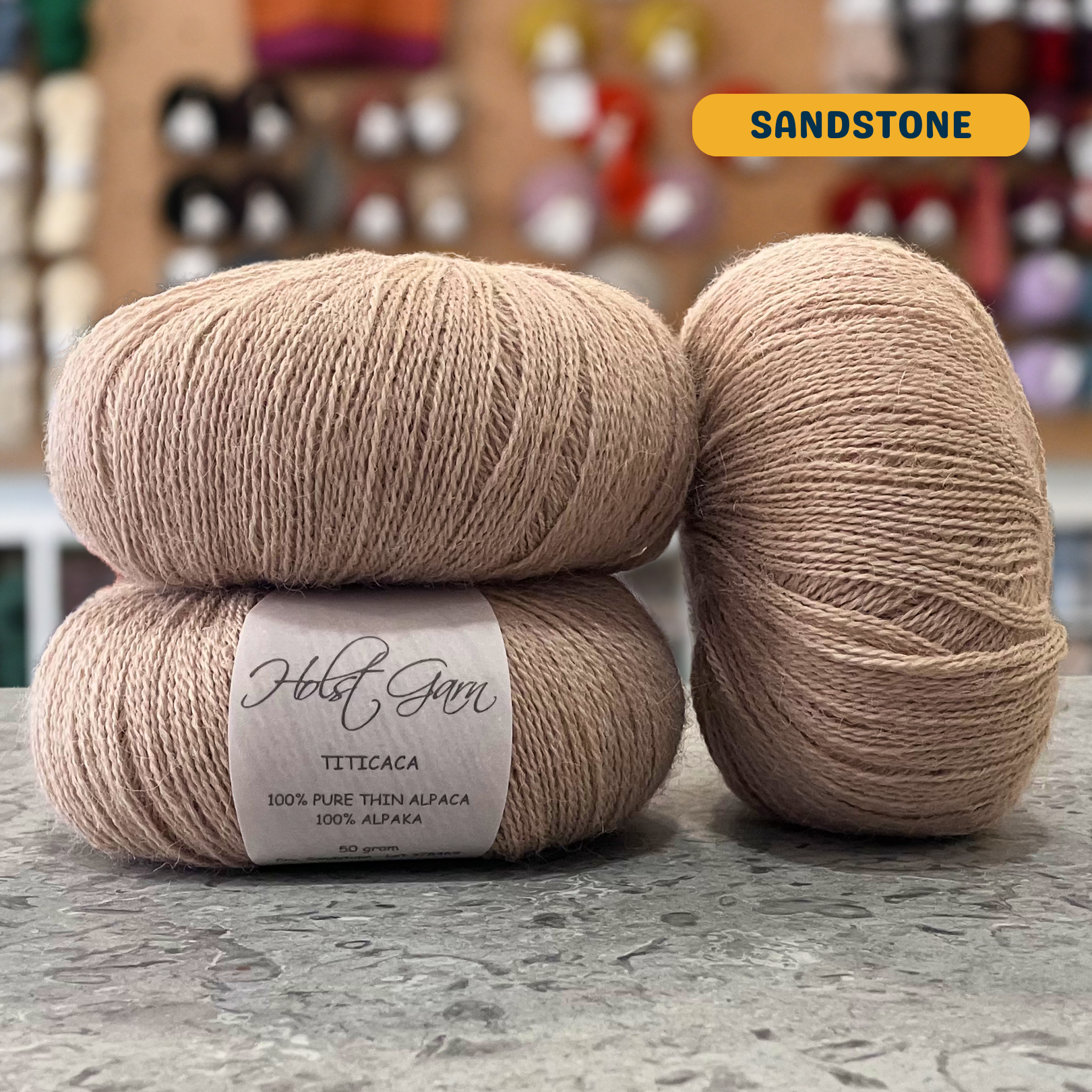 50g beige (sandstone) alpaca lace yarn, 'Holst Garn Titicaca 100% Pure Thin Alpaca' label visible, craft shop background (blurred)