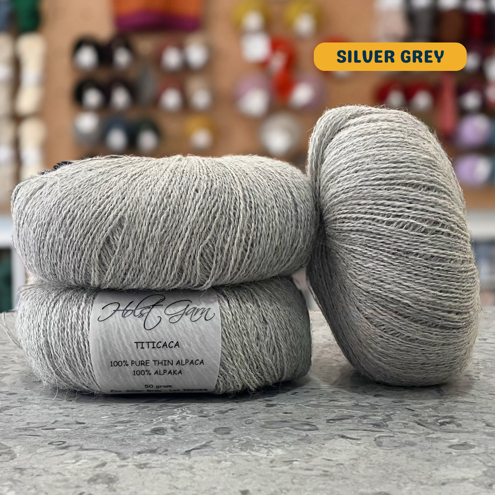 50g light grey (silver grey) alpaca lace yarn, 'Holst Garn Titicaca 100% Pure Thin Alpaca' label visible, craft shop background (blurred)