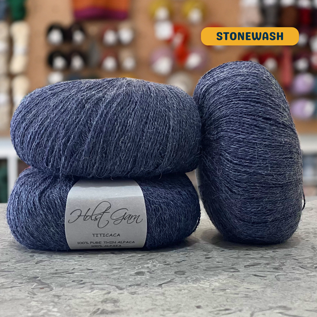50g dark blue-grey (stonewash) alpaca lace yarn, 'Holst Garn Titicaca 100% Pure Thin Alpaca' label visible, craft shop background (blurred)