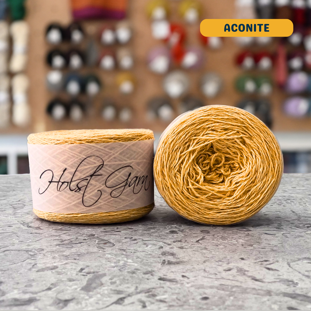 50g yellow (aconite) lambswool‑merino‑cotton light fingering skein, 'Holst Garn' label visible, craft shop background (blurred)