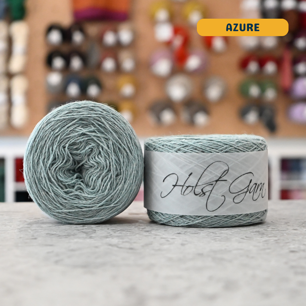 50g light azure lambswool‑merino‑cotton light fingering skein, 'Holst Garn' label visible, craft shop background (blurred)