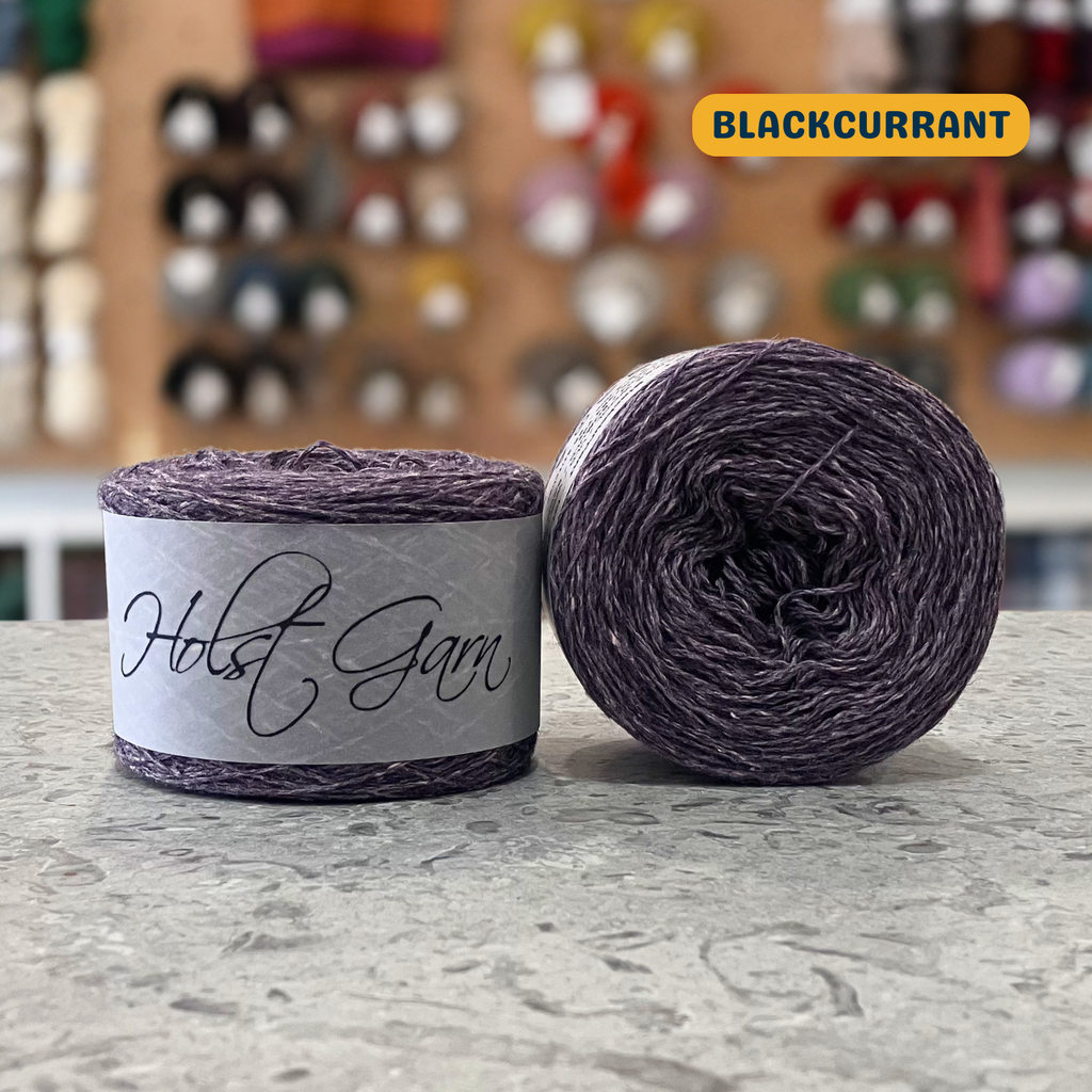 50g grey-purple (blackcurrant) lambswool‑merino‑cotton light fingering skein, 'Holst Garn' label visible, craft shop background (blurred)