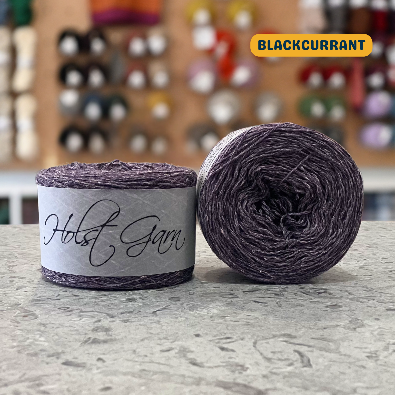 50g grey-purple (blackcurrant) lambswool‑merino‑cotton light fingering skein, 'Holst Garn' label visible, craft shop background (blurred)