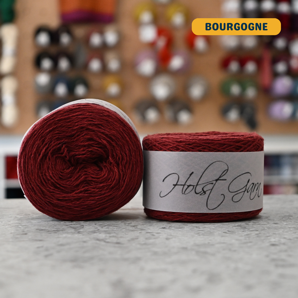 50g deep red (bourgogne) lambswool‑merino‑cotton light fingering skein, 'Holst Garn' label visible, craft shop background (blurred)