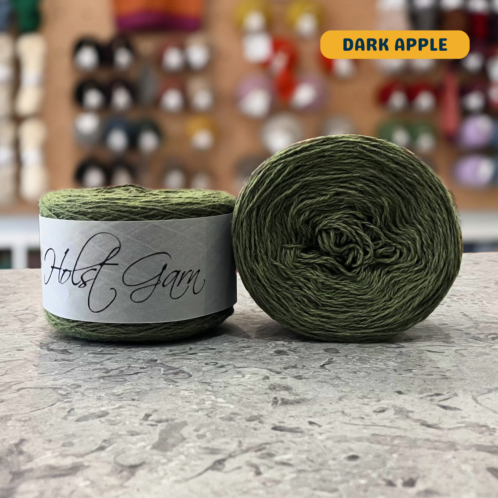 50g green (dark apple) lambswool‑merino‑cotton light fingering skein, 'Holst Garn' label visible, craft shop background (blurred)