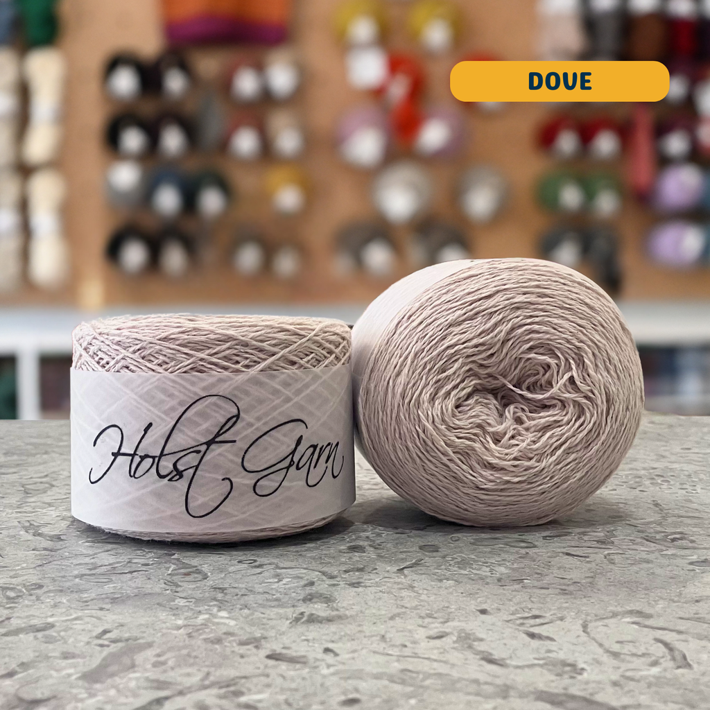 50g beige (dove) lambswool‑merino‑cotton light  fingering skein, 'Holst Garn' label visible, craft shop background (blurred)