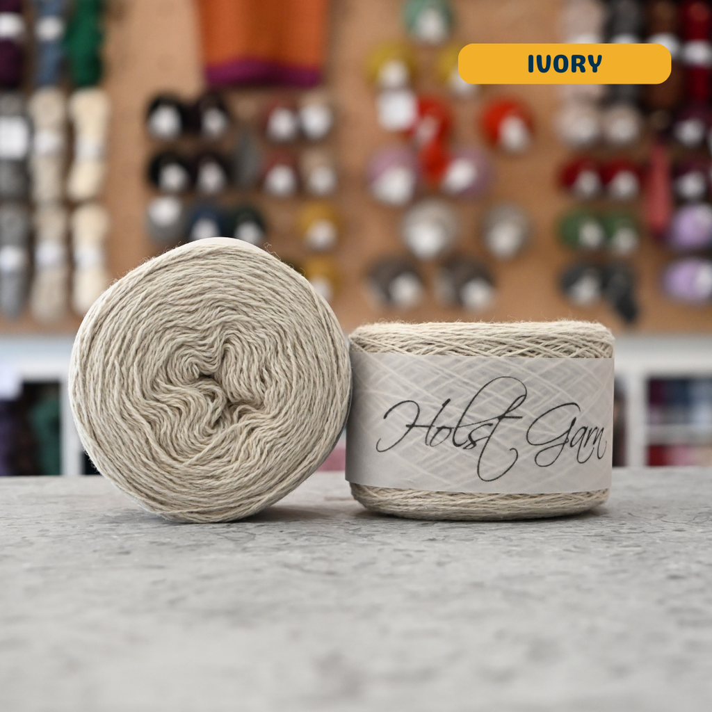 50g ivory lambswool‑merino‑cotton light fingering skein, 'Holst Garn' label visible, craft shop background (blurred)