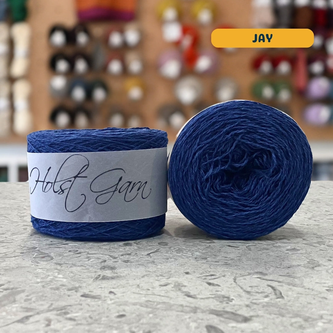 50g dark blue (jay) lambswool‑merino‑cotton light fingering skein, 'Holst Garn' label visible, craft shop background (blurred)