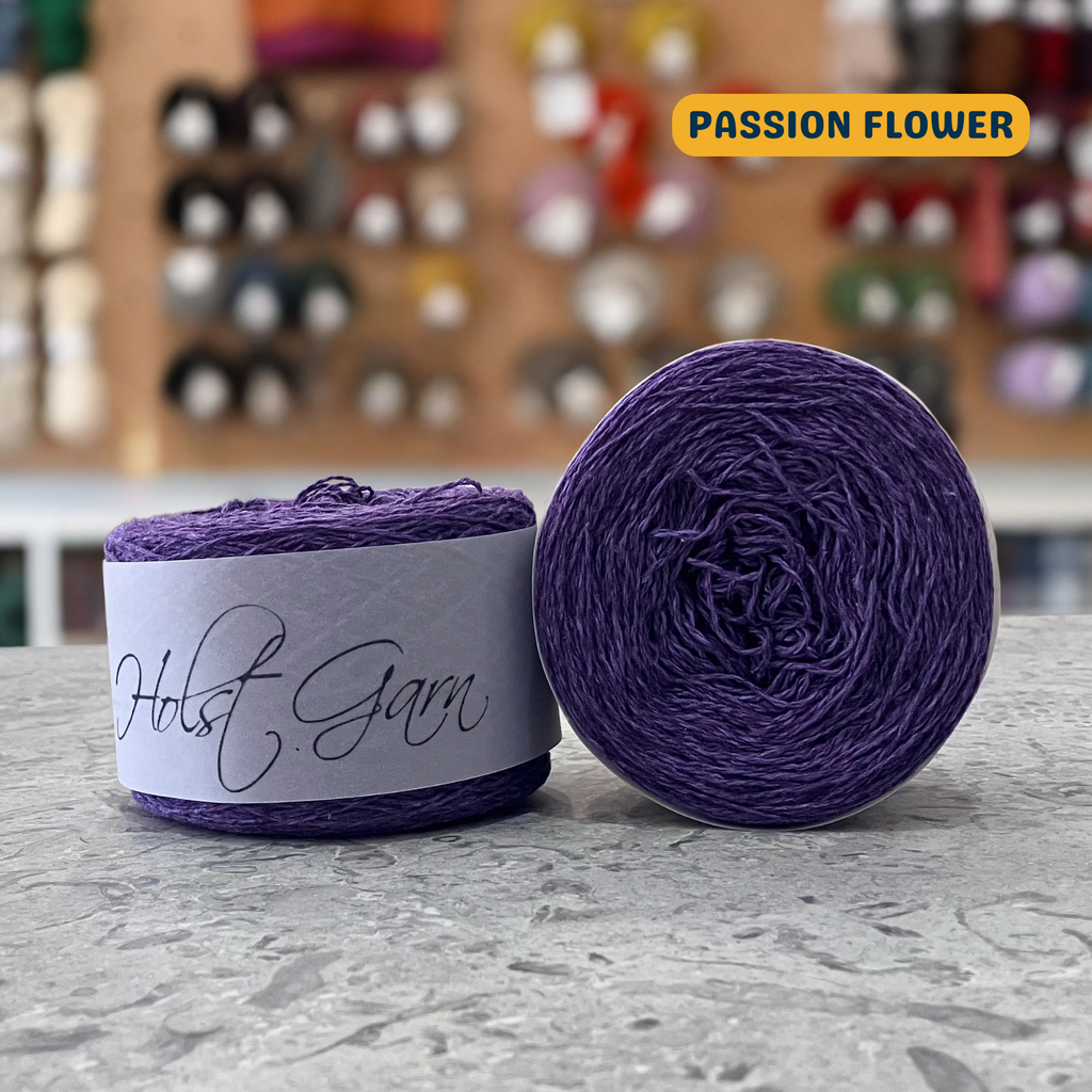 50g purple (passion flower) lambswool‑merino‑cotton light fingering skein, 'Holst Garn' label visible, craft shop background (blurred)