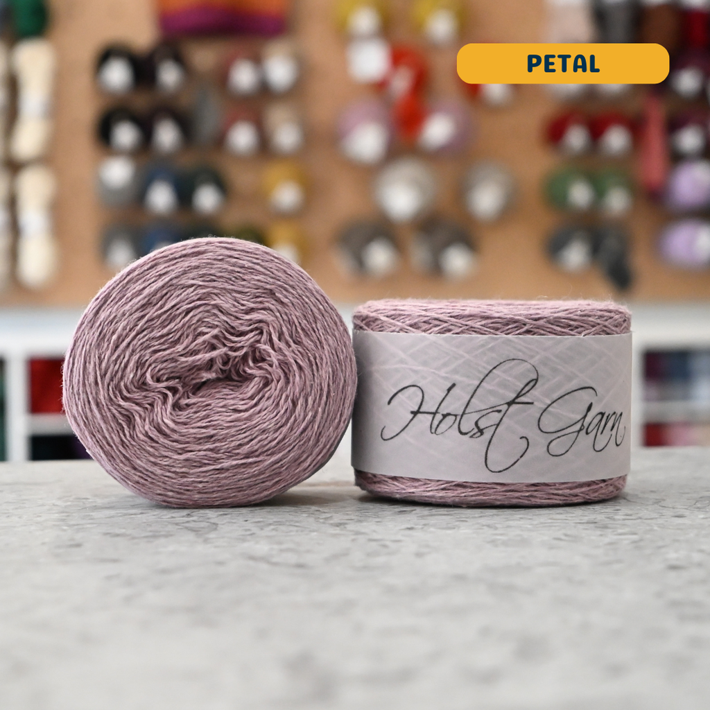 50g pale pink (petal) lambswool‑merino‑cotton light fingering skein, 'Holst Garn' label visible, craft shop background (blurred)