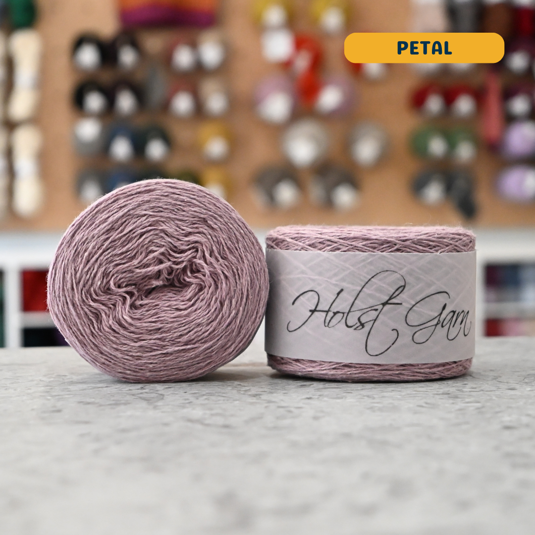 50g pale pink (petal) lambswool‑merino‑cotton light fingering skein, 'Holst Garn' label visible, craft shop background (blurred)