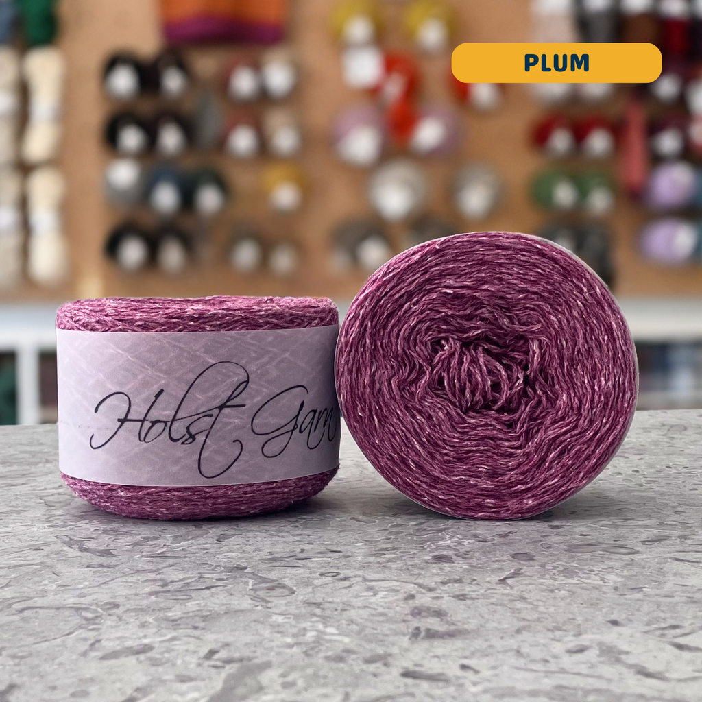 50g pink-purple (plum) lambswool‑merino‑cotton light fingering skein, 'Holst Garn' label visible, craft shop background (blurred)