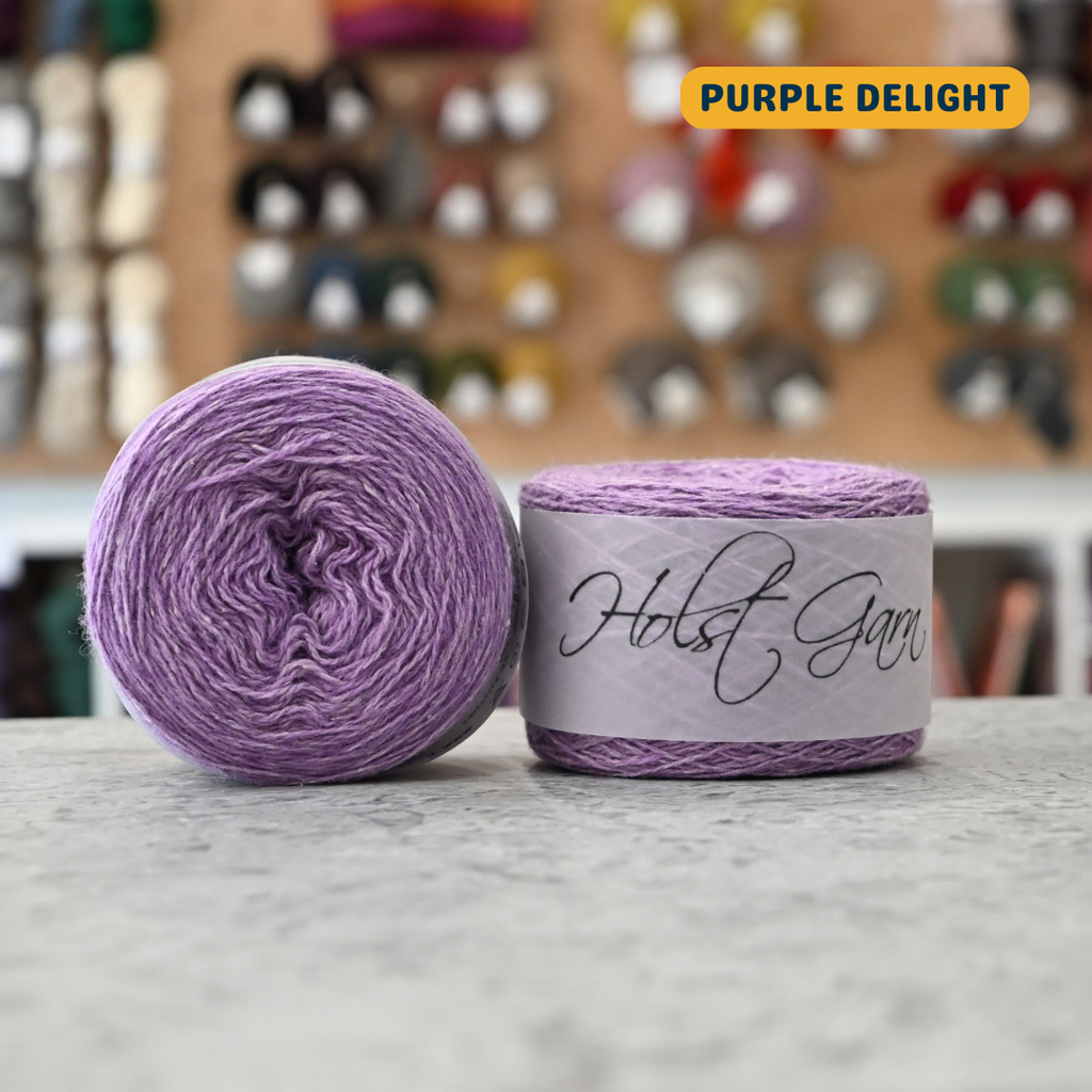 50g lilac (purple delight) lambswool‑merino‑cotton light fingering skein, 'Holst Garn' label visible, craft shop background (blurred)