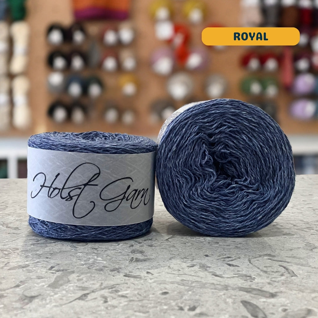 50g royal blue lambswool‑merino‑cotton light fingering skein, 'Holst Garn' label visible, craft shop background (blurred)
